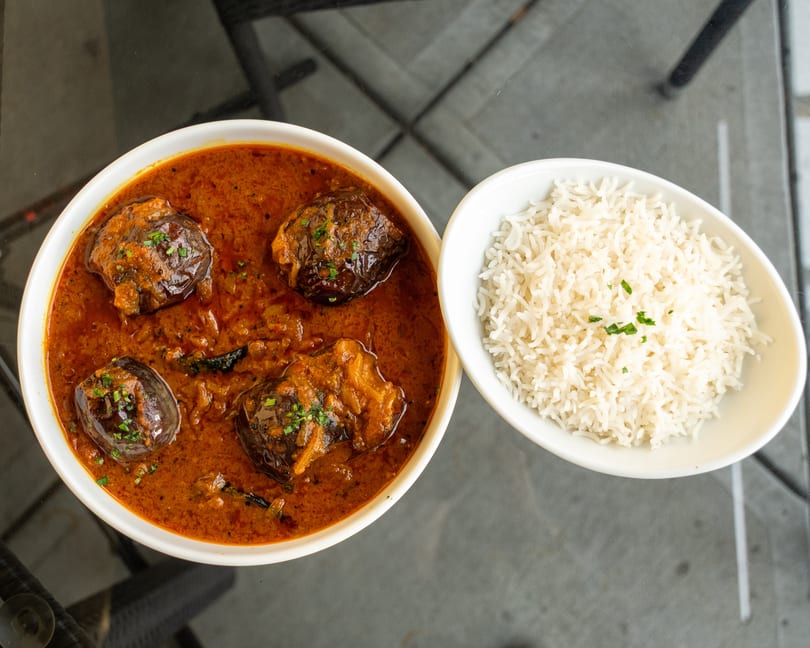 Eggplant curry in a white bowl next to a bowl of white rice, garnished with herbs.