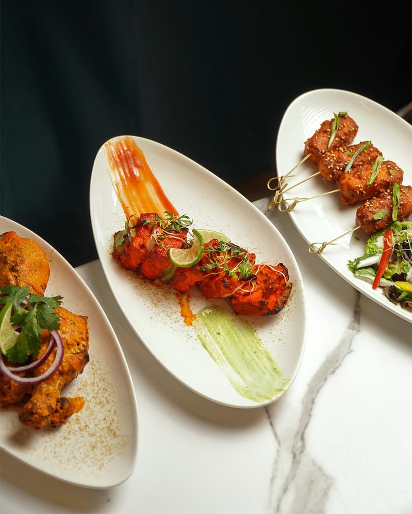 Three plates of vibrant Indian appetizers, including tandoori chicken and paneer skewers, on a marble table.