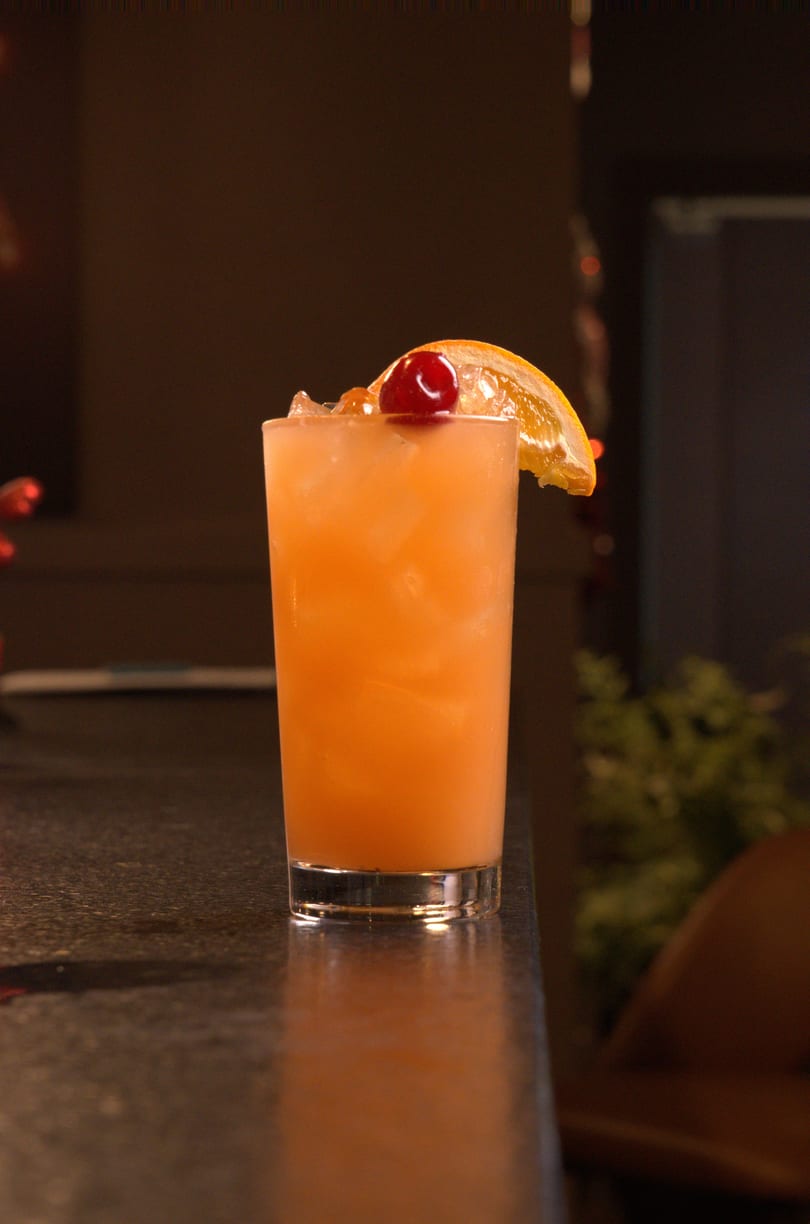 A vibrant orange cocktail with ice, garnished with a cherry and an orange slice on a dark bar.