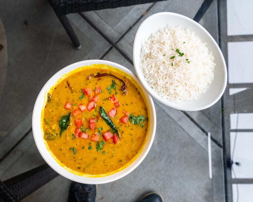 Overhead view of a bowl of yellow dal with tomatoes and herbs, next to white rice.