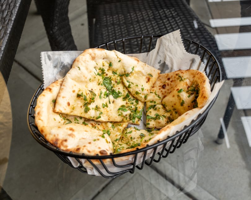 A black wire basket filled with warm, garlic naan bread, garnished with fresh parsley.