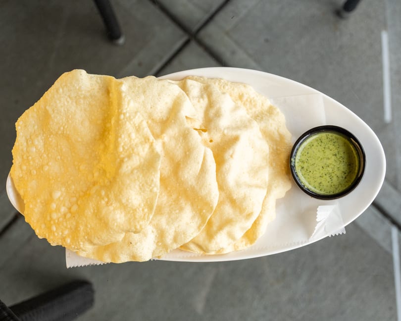 Crispy papadum crackers and a small bowl of green chutney on a white plate.