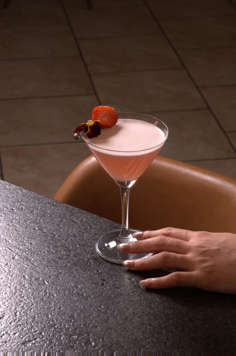A pink cocktail in a martini glass with a strawberry and flower garnish, next to a person's hand.