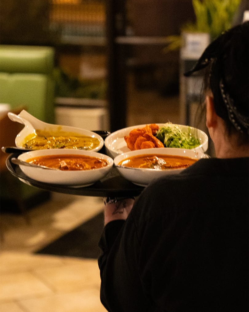 Server carrying a tray with four bowls of colorful Indian dishes through a restaurant.