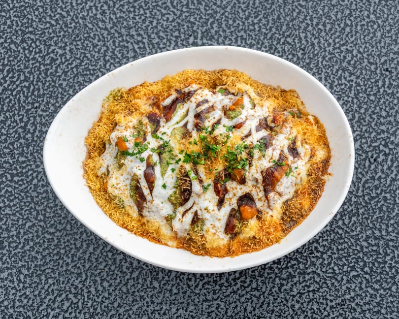 Overhead view of a white bowl filled with Indian chaat, topped with sauces and herbs.