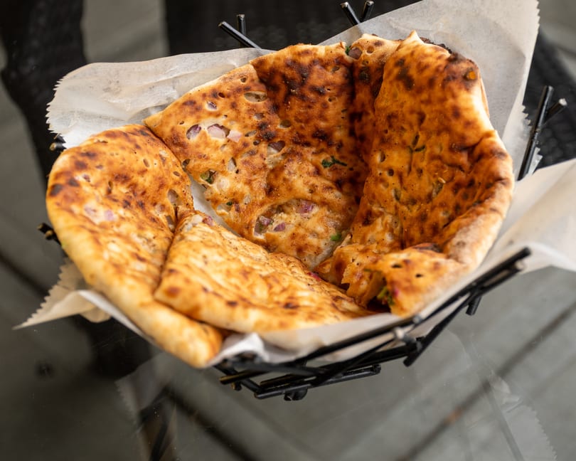 A basket of golden-brown naan bread, speckled with red onion and herbs, sits on a glass table.