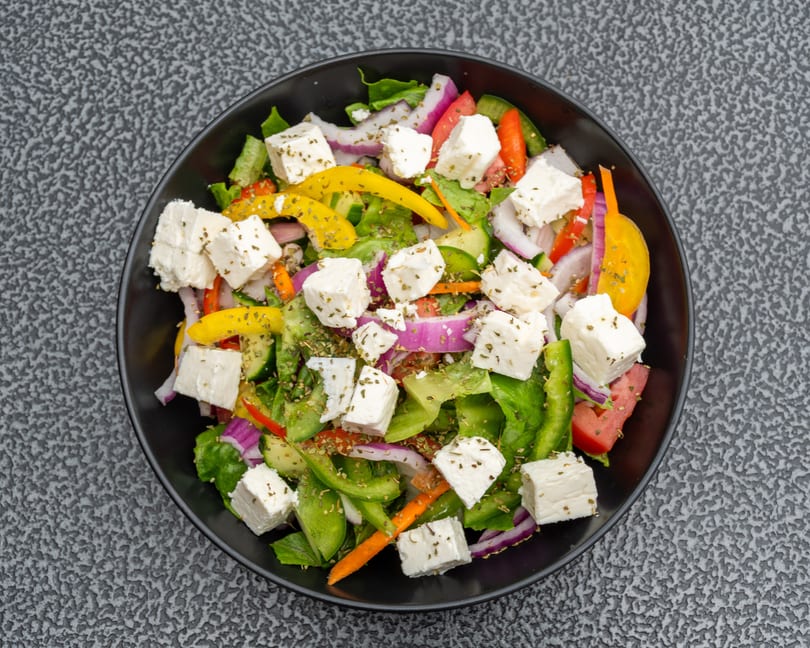 Overhead view of a vibrant Greek salad with feta, bell peppers, red onion, and lettuce in a black bowl.