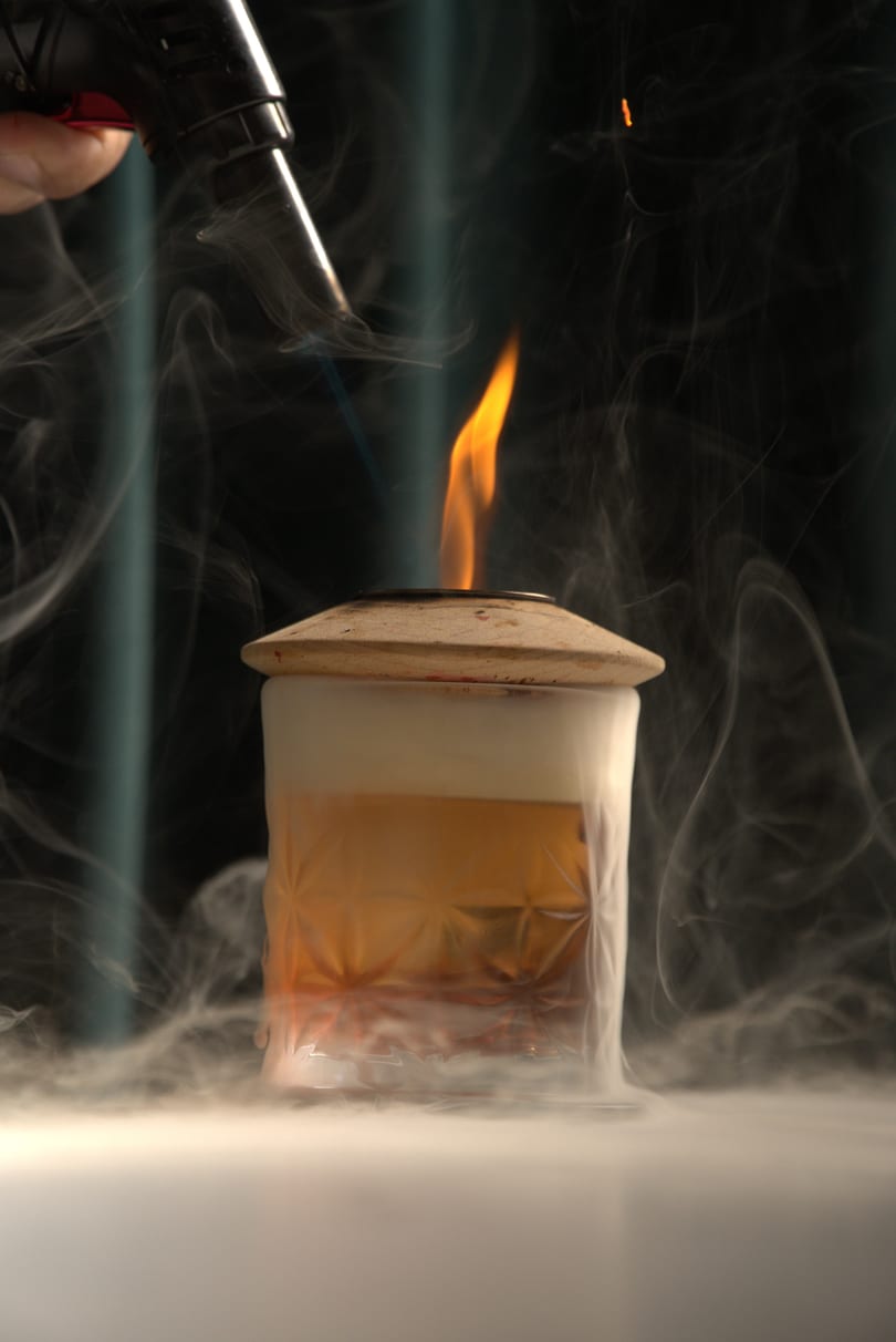 A cocktail in a patterned glass is being smoked with a torch, creating a fiery, misty effect.